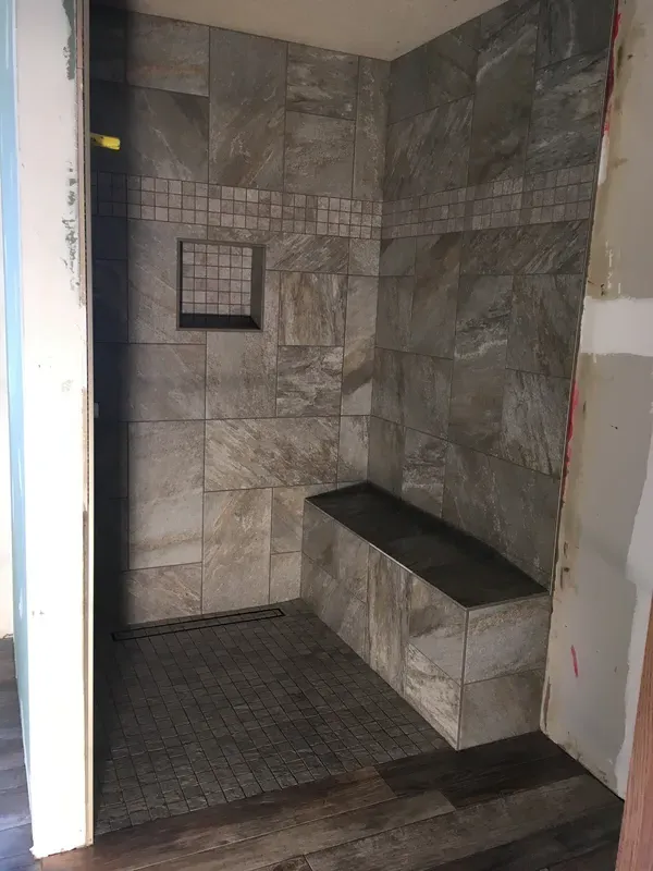 A gray walk in tile shower with stone seating and shower soap shelf